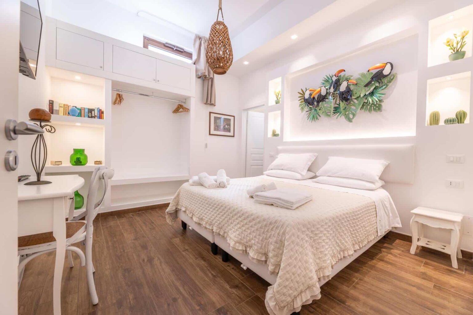 Cozy and modern bedroom with a white bedspread, decorative wall plants, and stylish wooden flooring. The room features a small desk, shelves with decorative items, and ample natural light, creating a welcoming atmosphere for relaxation. | Only on SopranoVillas