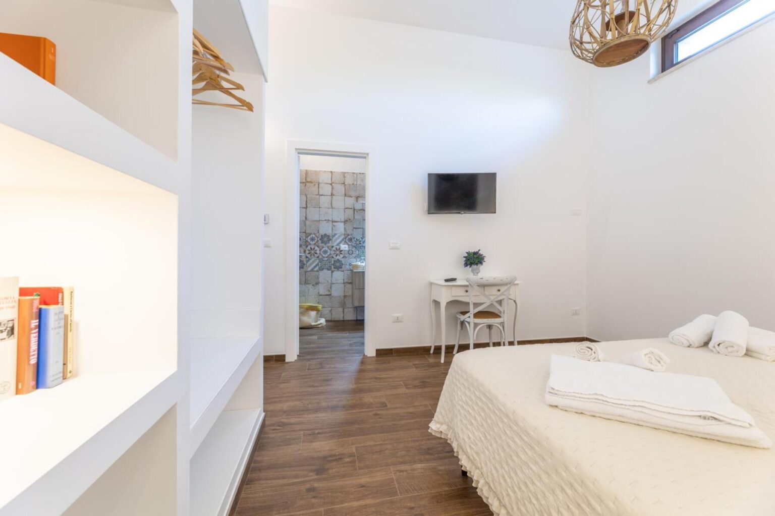 Modern bedroom with a queen-sized bed, featuring white bedding and neatly arranged towels. The room includes a small desk and chair, a wall-mounted TV, and a bookshelf with colorful books. A doorway leads to a stylish bathroom with tiled accents. Natural light streams in through a window, enhancing the inviting atmosphere. | Only on SopranoVillas