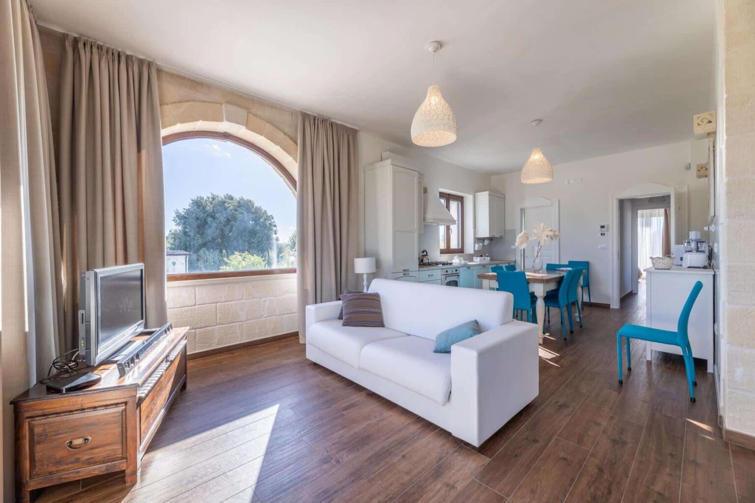 Bright and airy living room featuring a white sofa, wooden furniture, and large windows with curtains, showcasing a modern kitchen and blue dining chairs in the background. Ideal for showcasing contemporary interior design. | Only on SopranoVillas