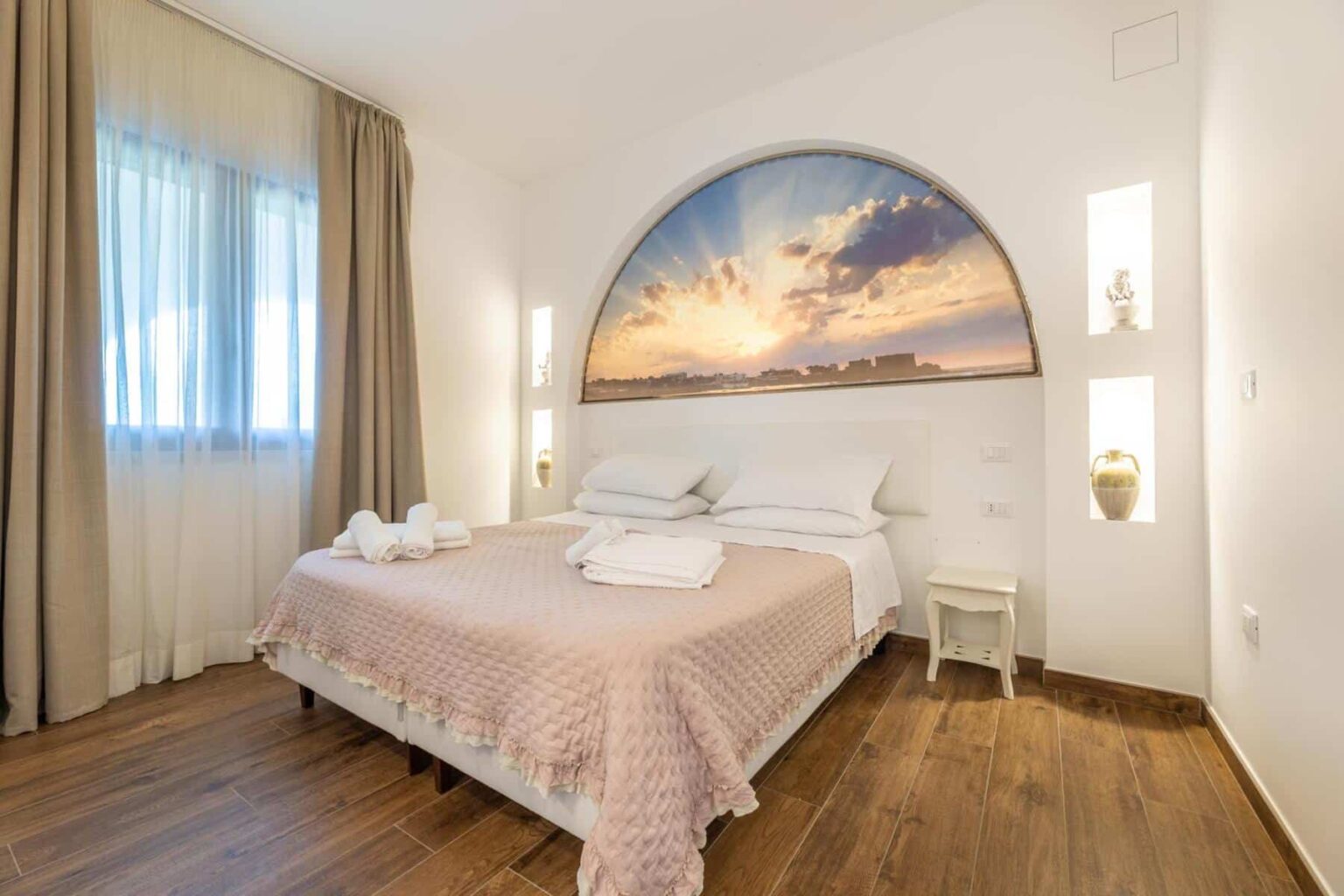 Cozy bedroom featuring a large bed with soft linens, a decorative sunset mural above the headboard, natural light from a window, and elegant wooden flooring. Ideal for relaxation and comfort. | Only on SopranoVillas