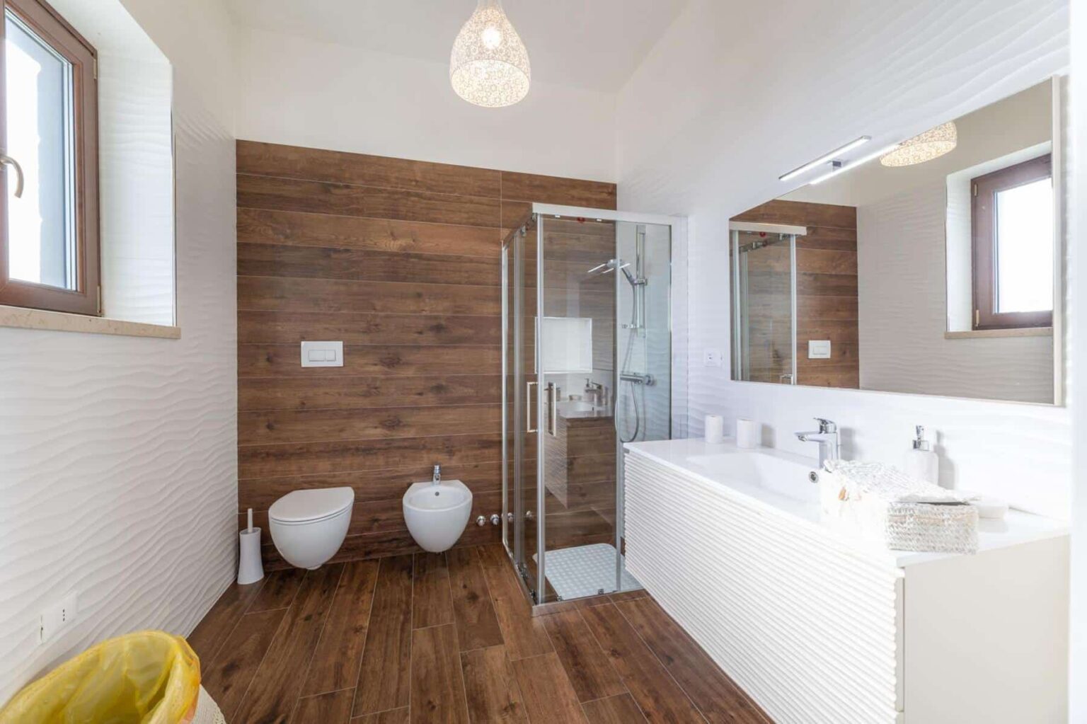 Modern bathroom interior featuring a glass shower, dual toilet and bidet, wooden accent wall, and elegant lighting. The sleek design includes a white vanity with a textured front and decorative accessories, showcasing a blend of functionality and style. | Only on SopranoVillas