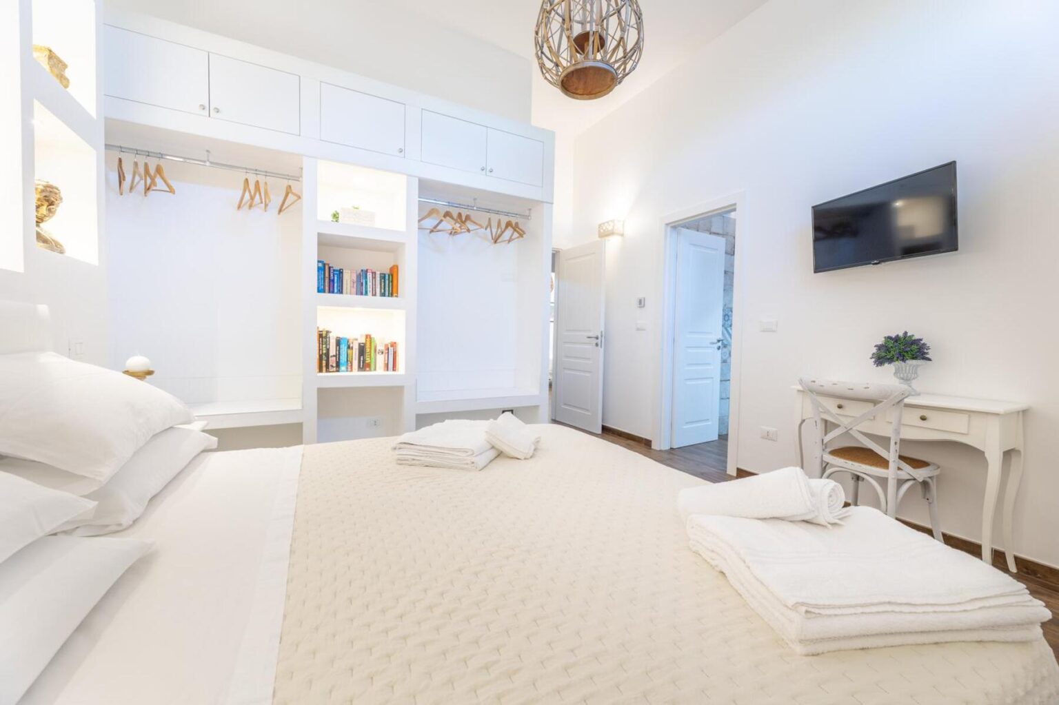 Modern and cozy bedroom featuring a comfortable bed with white linens, neatly arranged towels, a stylish desk, and a wall-mounted TV. The space includes built-in shelving with books and decorative items, along with a closet area with wooden hangers. Natural light enhances the bright, minimalist decor, creating a welcoming atmosphere. | Only on SopranoVillas