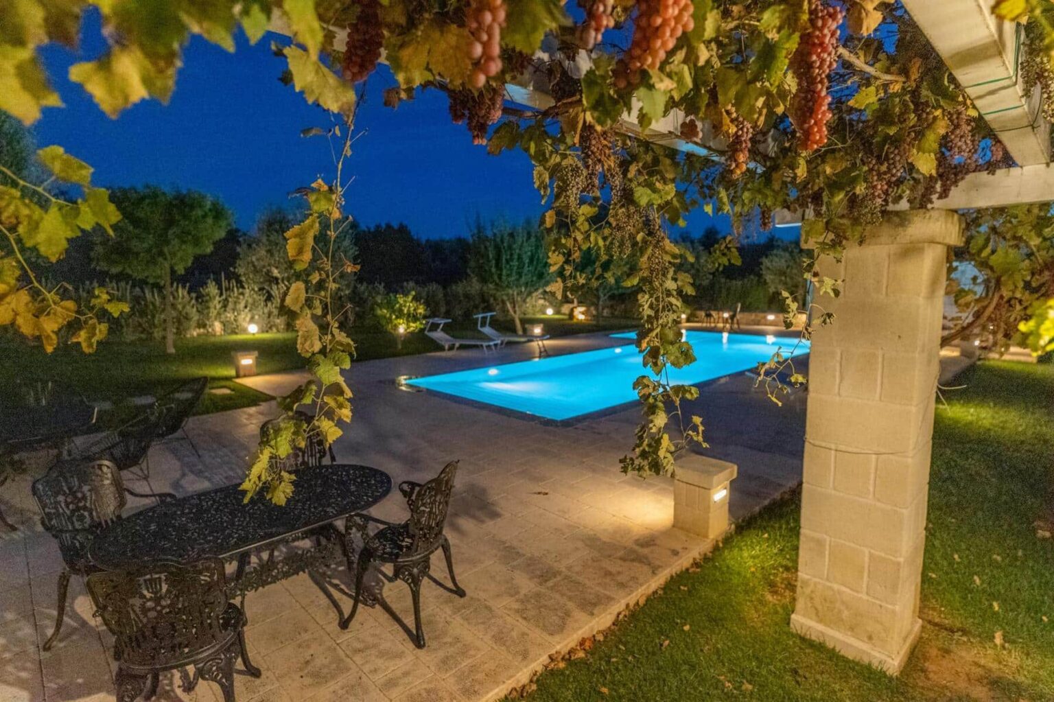 Beautiful outdoor pool area at night, featuring a well-lit swimming pool surrounded by lush greenery and grapevines. Elegant black wrought iron furniture is set up for relaxation, creating a serene atmosphere perfect for evening gatherings. | Only on SopranoVillas