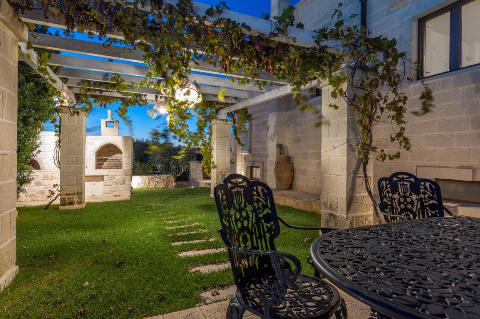 Charming outdoor patio featuring a stone structure with a vine-covered pergola, a dining table with ornate metal chairs, and a well-maintained lawn, creating an inviting space for relaxation and gatherings. | Only on SopranoVillas