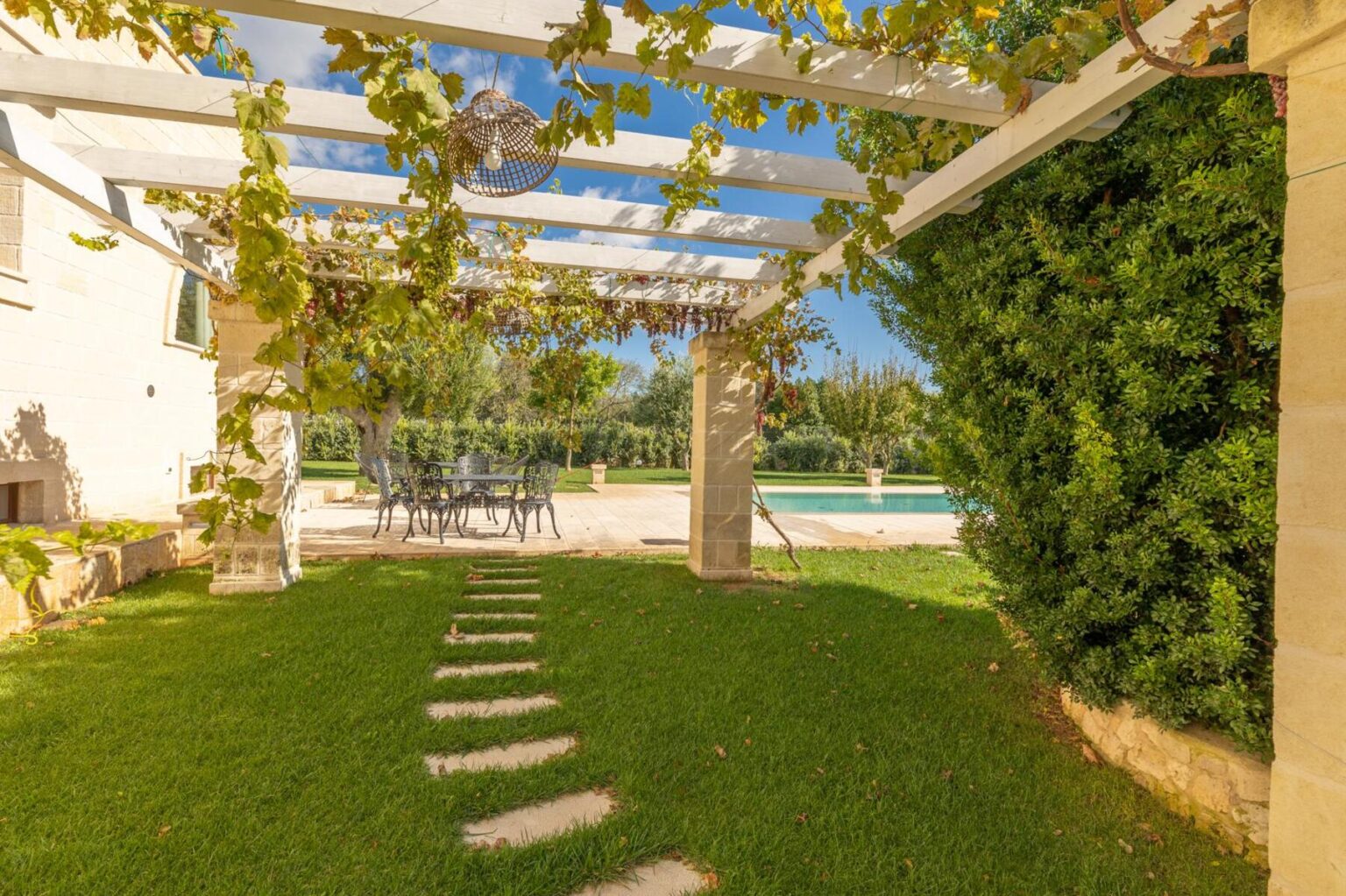 Beautiful outdoor patio with a vine-covered pergola, featuring a stone pathway leading to a swimming pool, surrounded by lush greenery and a dining set for al fresco meals. Ideal for relaxation and entertaining in a serene garden setting. | Only on SopranoVillas