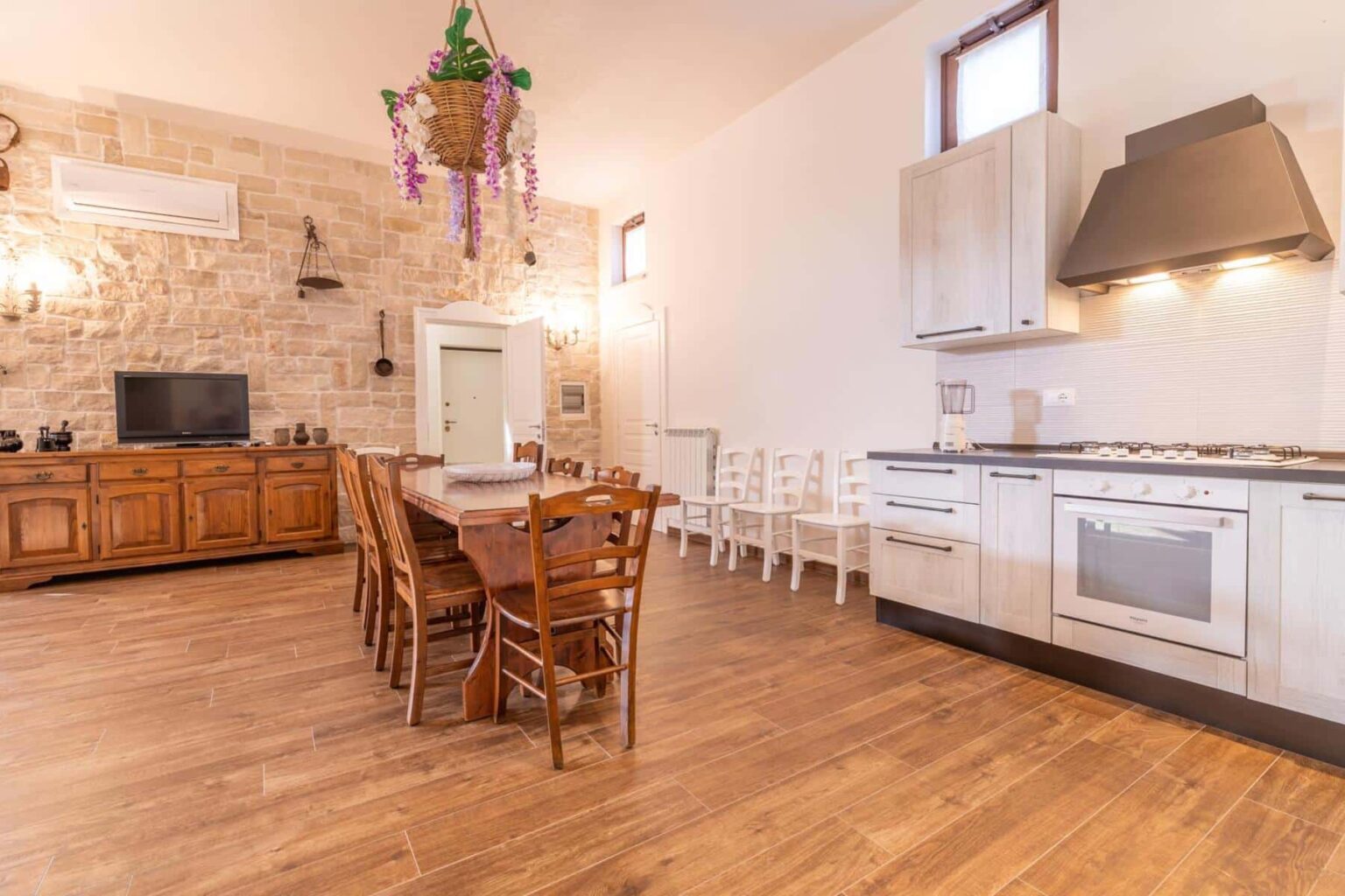 Modern kitchen and dining area featuring wooden furniture, a stone accent wall, and contemporary appliances. The space includes a large dining table with wooden chairs, a sleek kitchen with a stove and hood, and decorative plants hanging from the ceiling, creating a warm and inviting atmosphere. | Only on SopranoVillas
