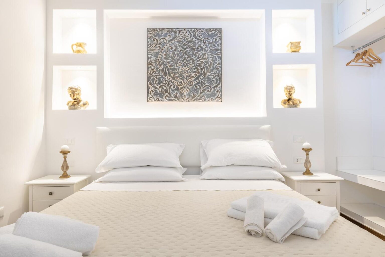 Cozy and stylish bedroom featuring a queen bed with white linens, decorative pillows, and a soft beige quilt. The room is illuminated by built-in shelves displaying elegant sculptures and a decorative wall art piece, enhancing the modern aesthetic. White walls and minimalist furniture create a bright and inviting atmosphere. | Only on SopranoVillas