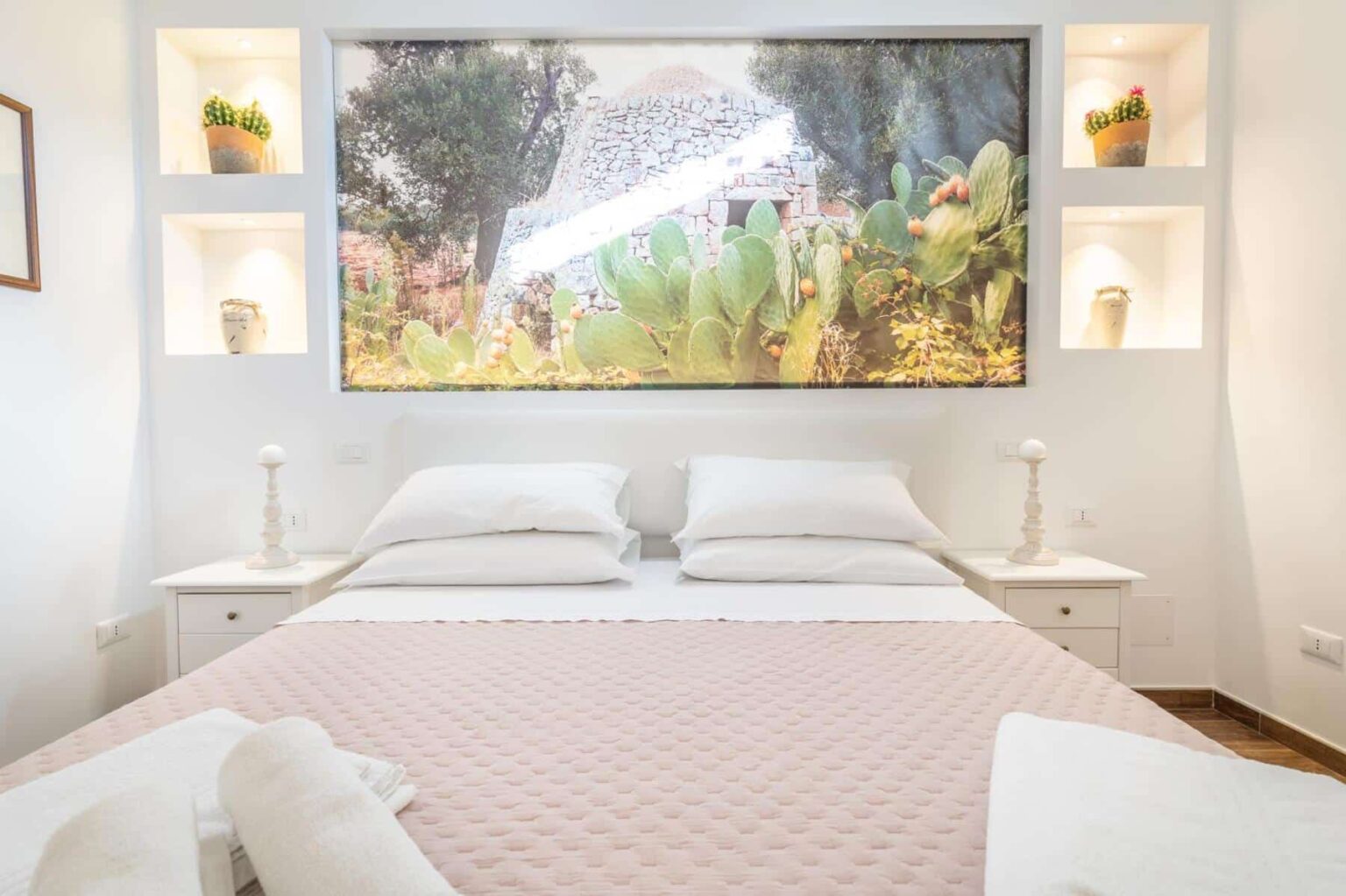 Cozy bedroom featuring a comfortable double bed with white linens, a decorative pink throw, and a large framed landscape artwork above. The room is illuminated by soft lighting from built-in shelves with potted plants, creating a serene and inviting atmosphere. | Only on SopranoVillas