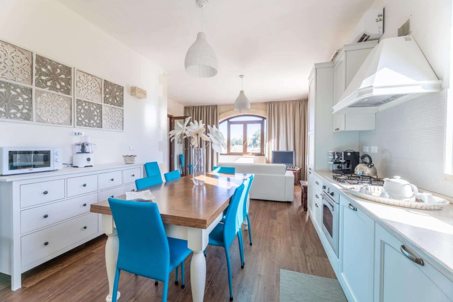Bright and modern kitchen with a wooden dining table surrounded by blue chairs, featuring white cabinetry, a microwave, and a cozy living area in the background. Natural light floods the space through large windows, enhancing the inviting atmosphere. | Only on SopranoVillas