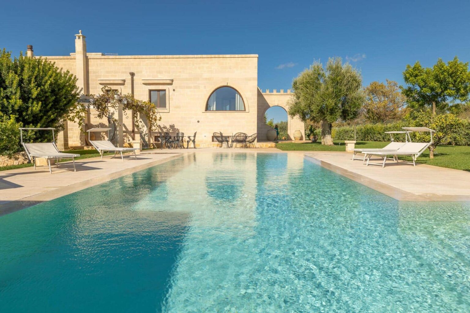 Luxurious villa with a serene swimming pool surrounded by lush greenery and comfortable lounge chairs, set against a clear blue sky. Ideal for relaxation and outdoor gatherings. | Only on SopranoVillas