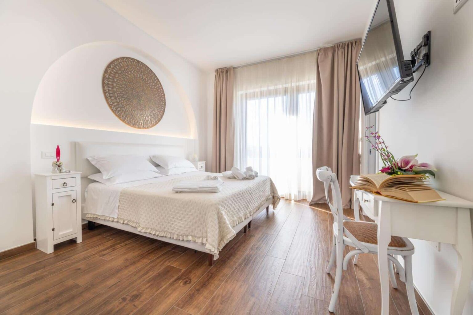Modern hotel room featuring a comfortable bed with white linens, a decorative wall art piece, and a flat-screen TV. The space includes a small writing desk with an open book and fresh flowers, large windows dressed with sheer curtains, and wooden flooring, creating a bright and inviting atmosphere. | Only on SopranoVillas