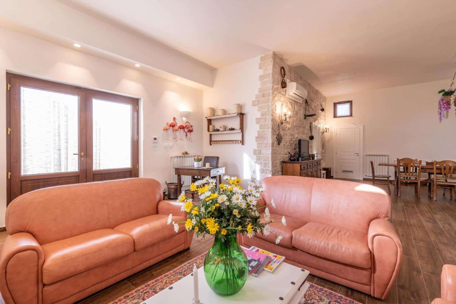 Cozy living room featuring two light brown leather sofas, a green vase with yellow and white flowers on a coffee table, and a rustic stone wall. Natural light streams through double doors, illuminating a wooden dining table and decorative elements like wall art and a television stand. | Only on SopranoVillas