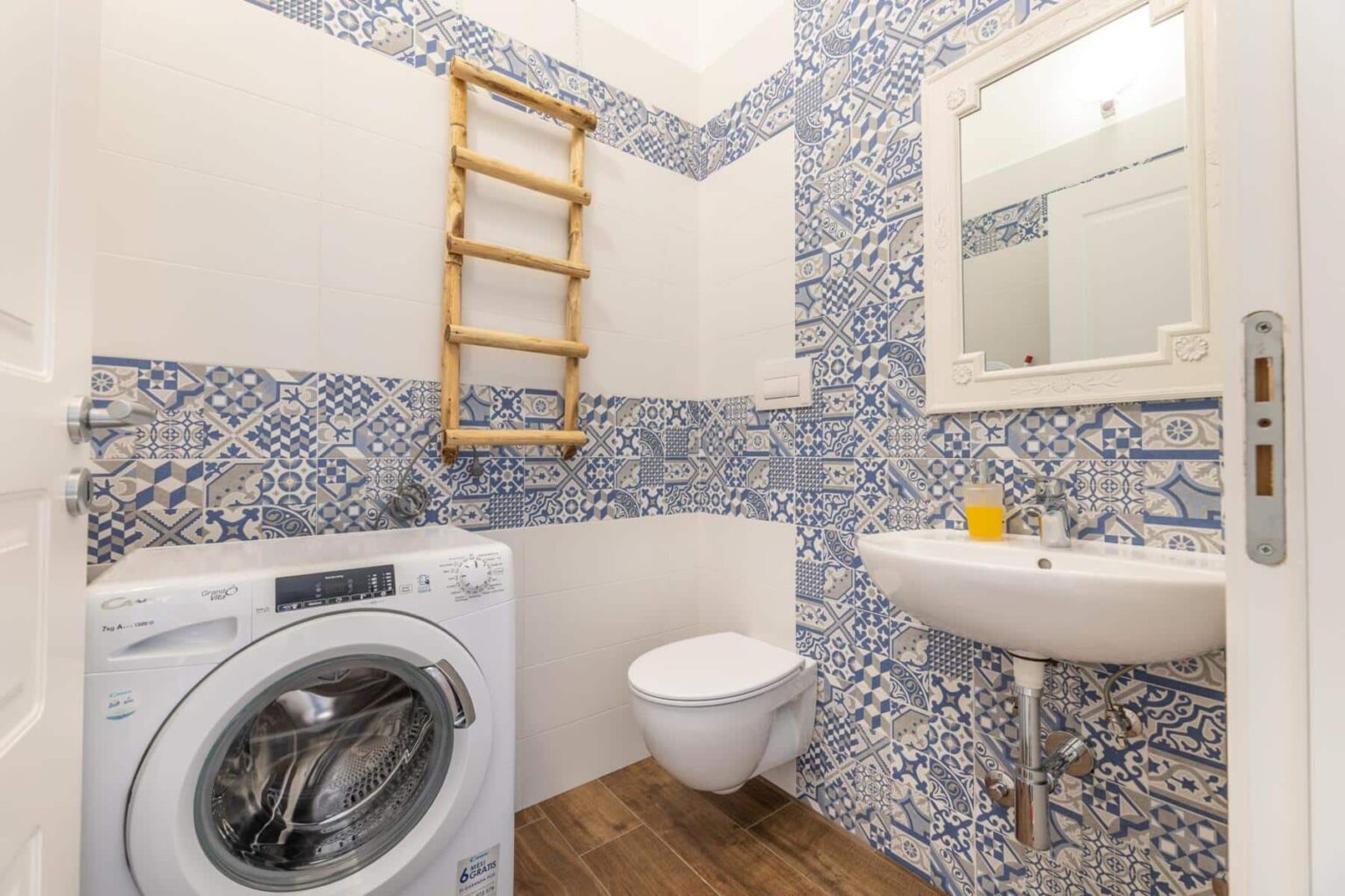 Modern bathroom with blue patterned tiles, featuring a washing machine, wall-mounted mirror, sink, and toilet. A wooden ladder adds decorative storage space. | Only on SopranoVillas