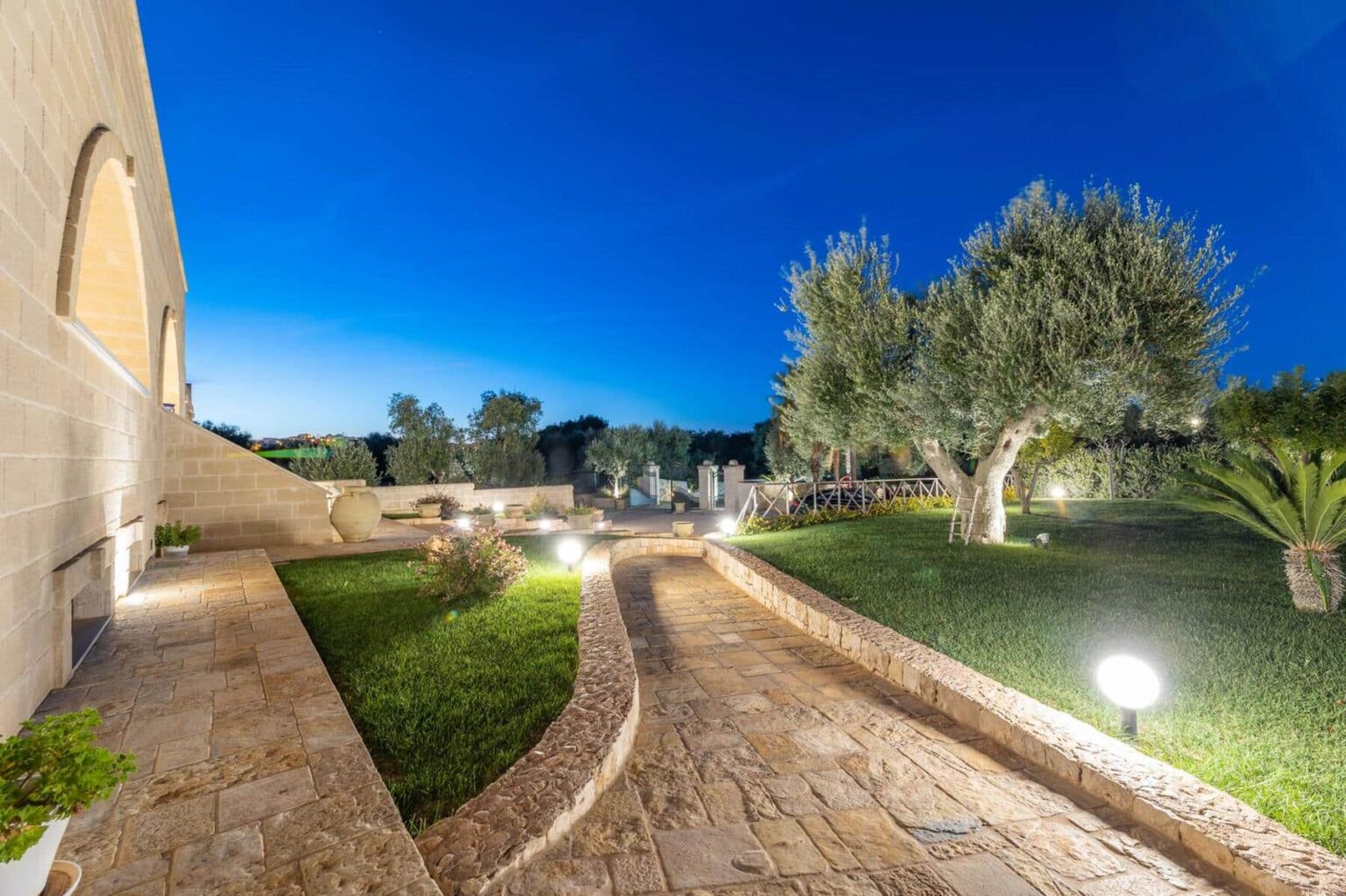 Pathway illuminated by garden lights leading through a lush green landscape with olive trees, under a twilight sky. Ideal for showcasing outdoor landscaping and evening ambiance in residential or commercial properties. | Only on SopranoVillas