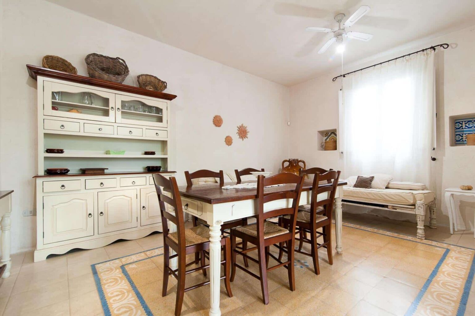 Bright and airy dining room featuring a wooden table with eight chairs, a classic white sideboard with glass display, and decorative wall elements. Natural light streams in through a large window with sheer curtains, enhancing the warm, inviting atmosphere. | Only on SopranoVillas