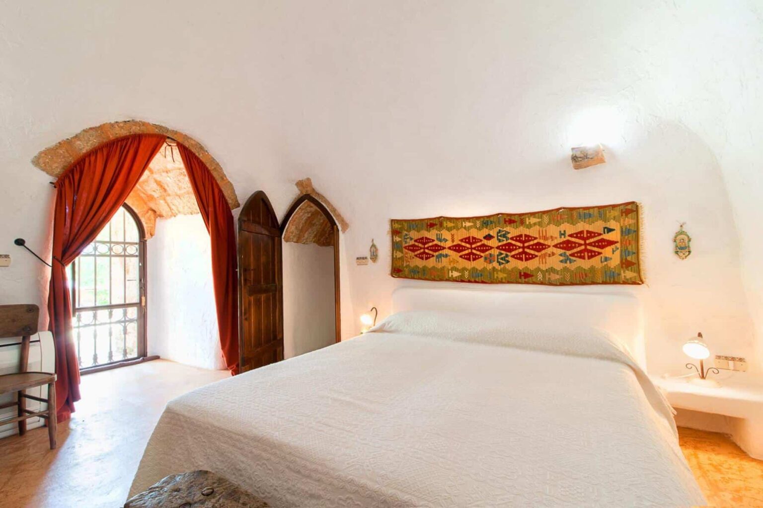 Cozy bedroom featuring a white bedspread, traditional decorative wall hanging, arched stone ceiling, and wooden accents. Natural light streams through a window with orange curtains, adding warmth to the space. | Only on SopranoVillas