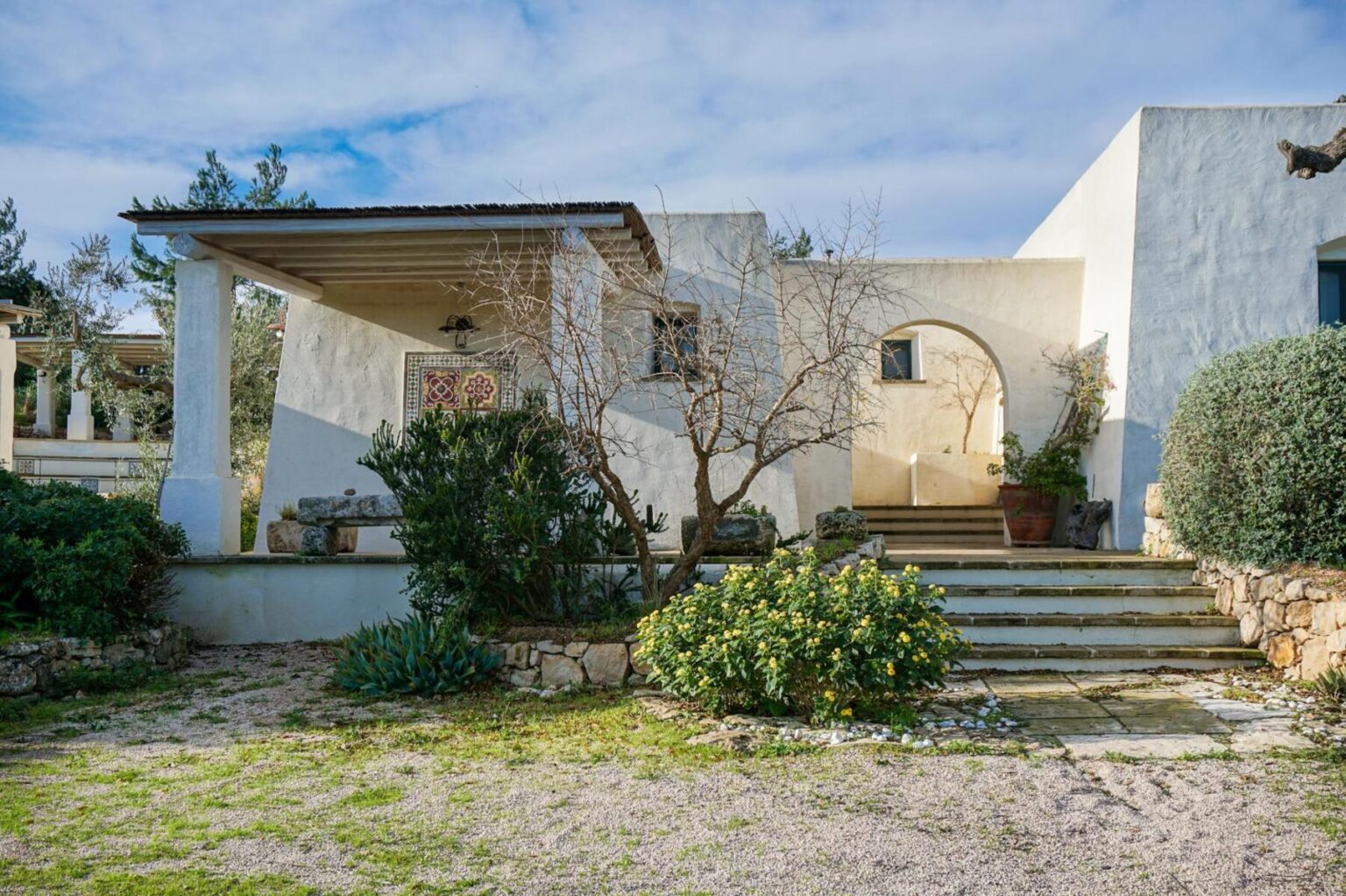 Charming white stucco house with a covered porch, decorative tile accents, and lush greenery in the foreground, set against a clear blue sky. Steps lead to an arched entrance, surrounded by colorful flowers and well-maintained landscaping. | Only on SopranoVillas