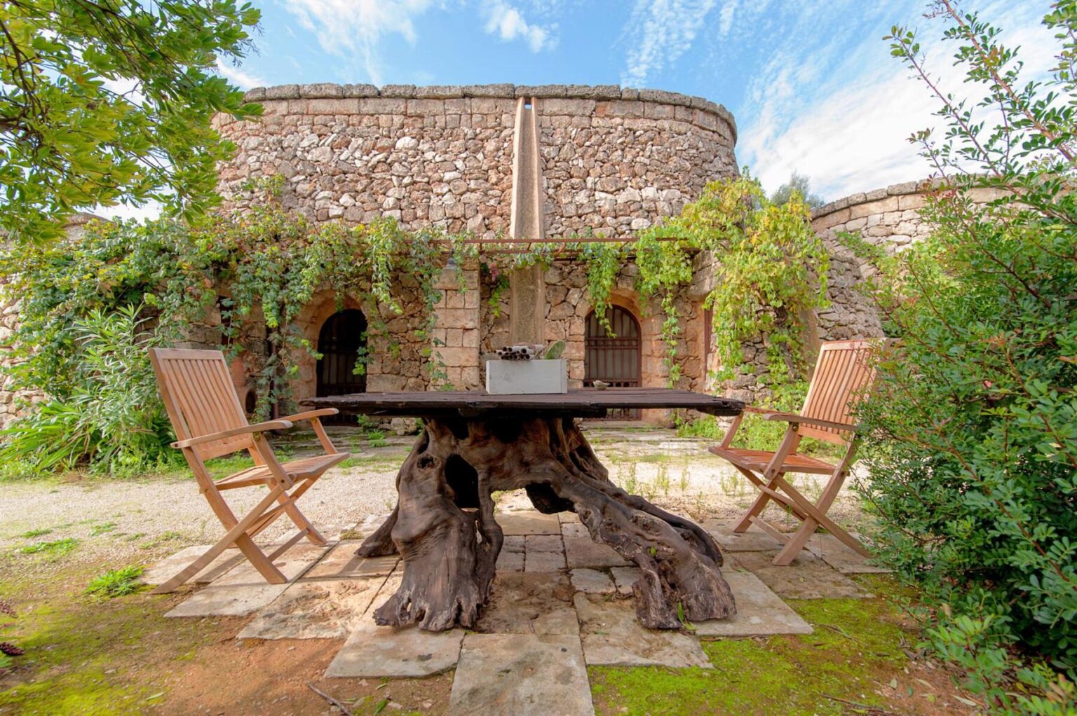Rustic outdoor dining area featuring a unique wooden table and chairs surrounded by lush greenery and a stone structure in the background. Ideal for nature lovers seeking a serene dining experience. | Only on SopranoVillas