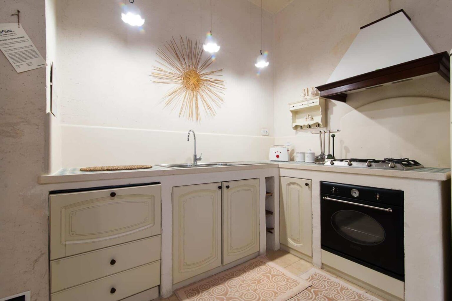 Modern kitchen with beige cabinets, stainless steel sink, and gas stove, featuring stylish overhead lighting and a decorative sunburst wall art. | Only on SopranoVillas