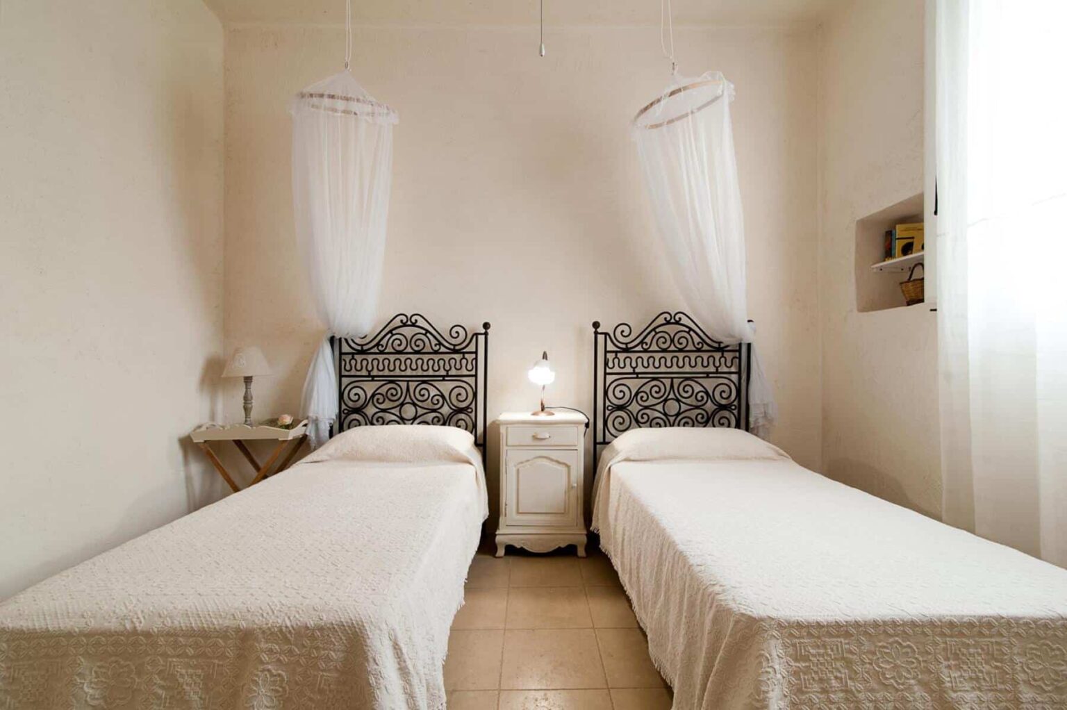 Cozy bedroom featuring two twin beds with ornate black metal headboards, soft white bedding, and a bedside table with a lamp, illuminated by natural light from a nearby window. Ideal for guest accommodations or family stays. | Only on SopranoVillas