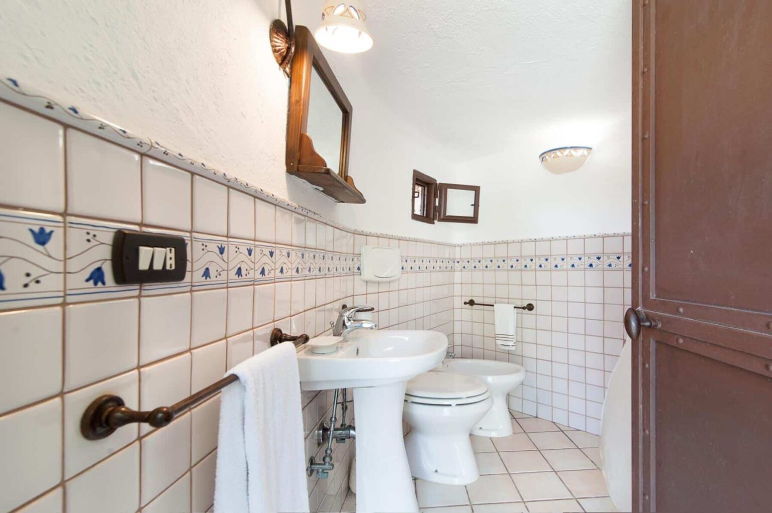 Bright and airy bathroom featuring white tiles, a pedestal sink, and a toilet, complemented by a wooden mirror and towel rack. The design includes decorative blue accents and natural light from a window. | Only on SopranoVillas
