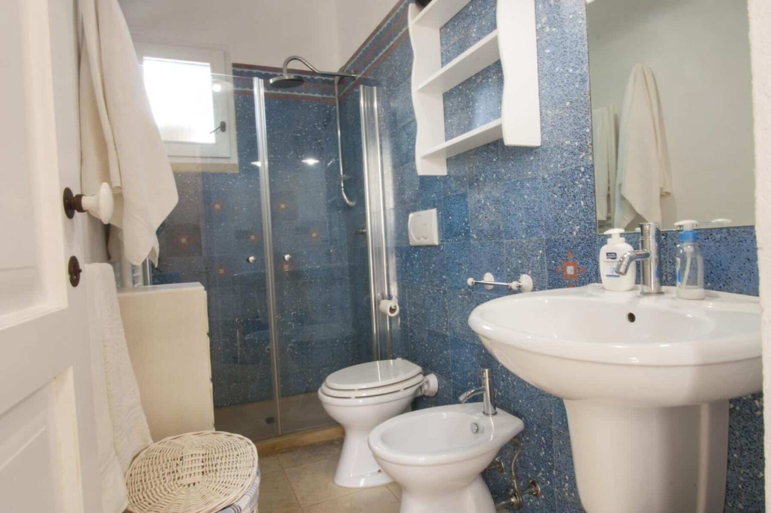 Modern bathroom featuring a glass shower, blue tiled walls, a pedestal sink, and a bidet, complemented by a white towel and decorative shelving. | Only on SopranoVillas