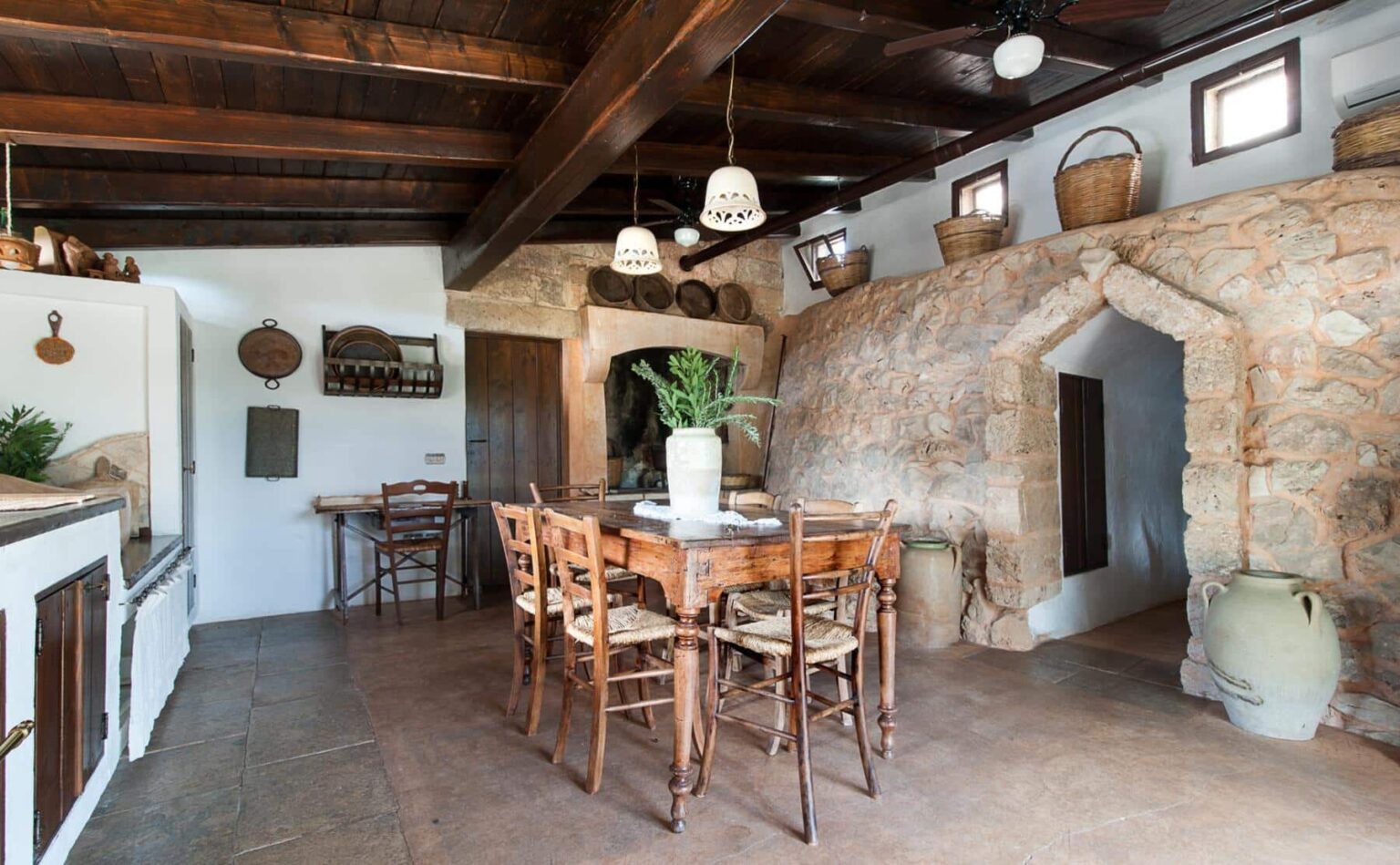 Rustic dining area featuring a wooden table surrounded by chairs, with a stone wall and traditional decor elements. The space includes wooden beams, potted plants, and decorative baskets, creating a warm and inviting atmosphere. | Only on SopranoVillas