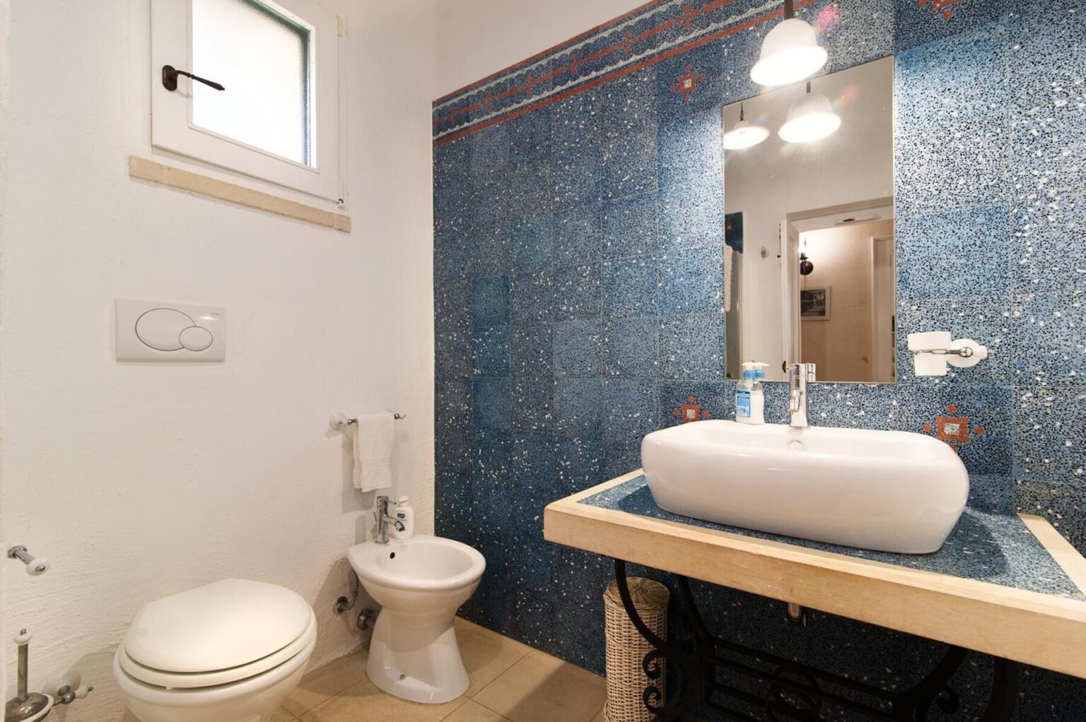 Modern bathroom featuring a blue mosaic wall, a stylish white vessel sink, a wooden countertop, a bidet, and a toilet, illuminated by warm overhead lighting. | Only on SopranoVillas