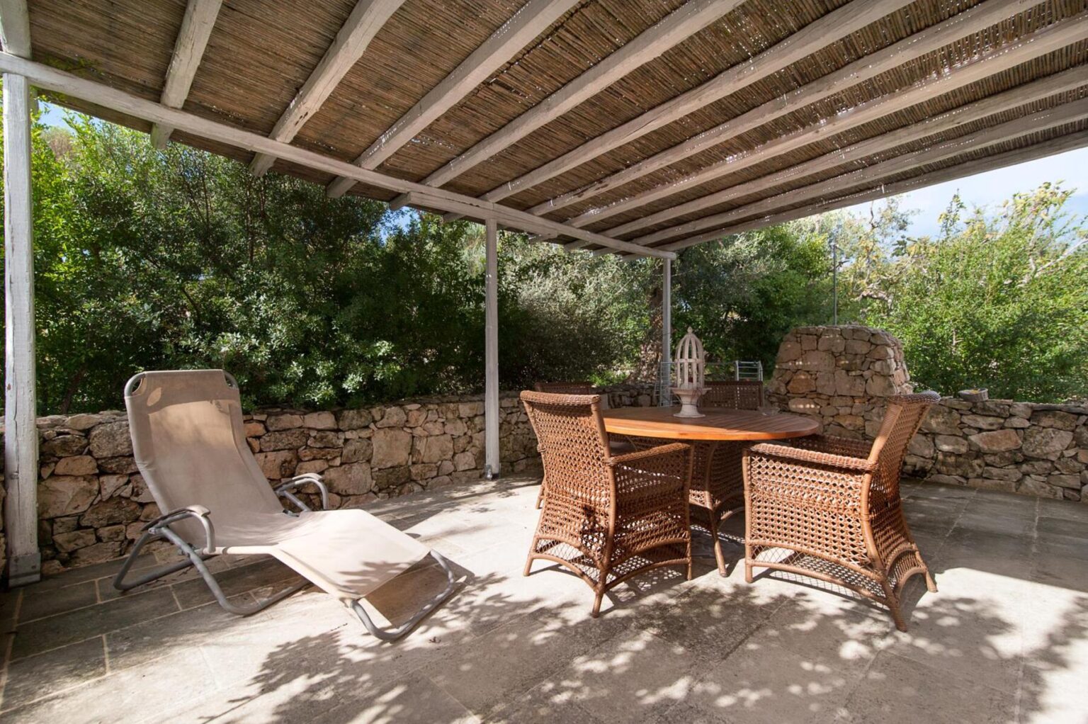Outdoor patio area featuring a wooden table surrounded by wicker chairs and a reclining lounge chair, with a stone wall and lush greenery in the background, providing a serene and inviting space for relaxation. | Only on SopranoVillas