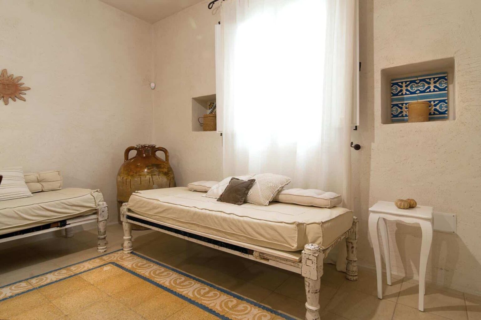 Bright and airy room featuring two elegant daybeds with neutral bedding, natural light streaming through a large window, and decorative wall niches showcasing intricate tile designs. The space has a calming atmosphere, enhanced by soft colors and minimalist decor. | Only on SopranoVillas