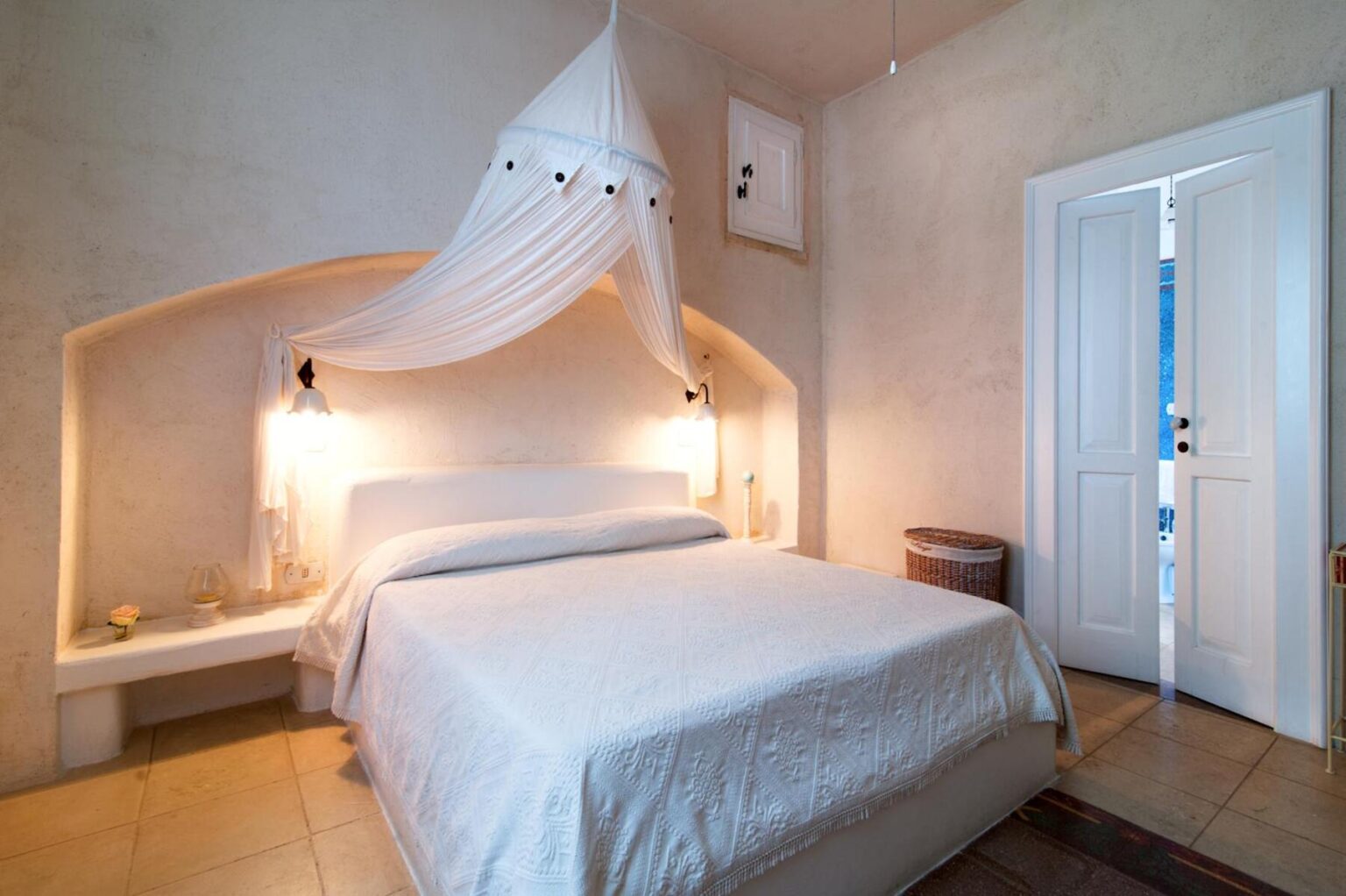 Cozy bedroom featuring a white bed with a canopy, soft lighting, and minimalist decor, complemented by an inviting doorway and natural light. Perfect for a serene retreat or vacation rental. | Only on SopranoVillas