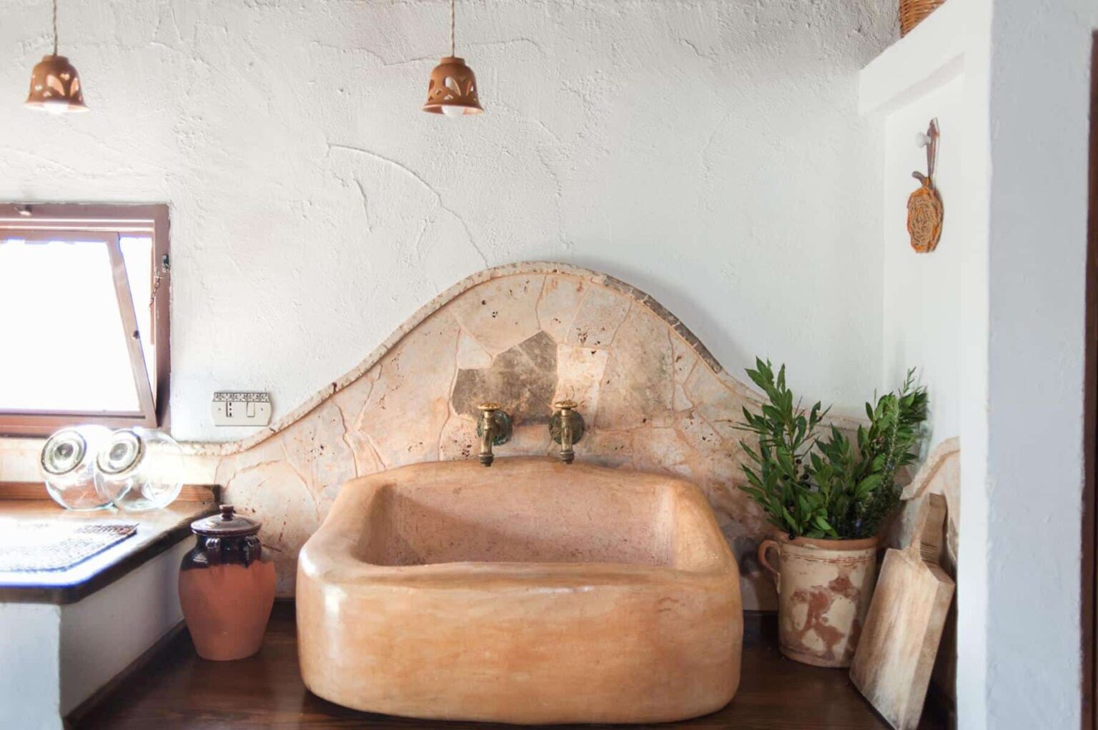 Rustic bathroom featuring a unique stone sink with vintage brass faucets, surrounded by natural decor including potted plants and traditional pottery. The space is illuminated by decorative pendant lights and has a warm, inviting atmosphere. | Only on SopranoVillas