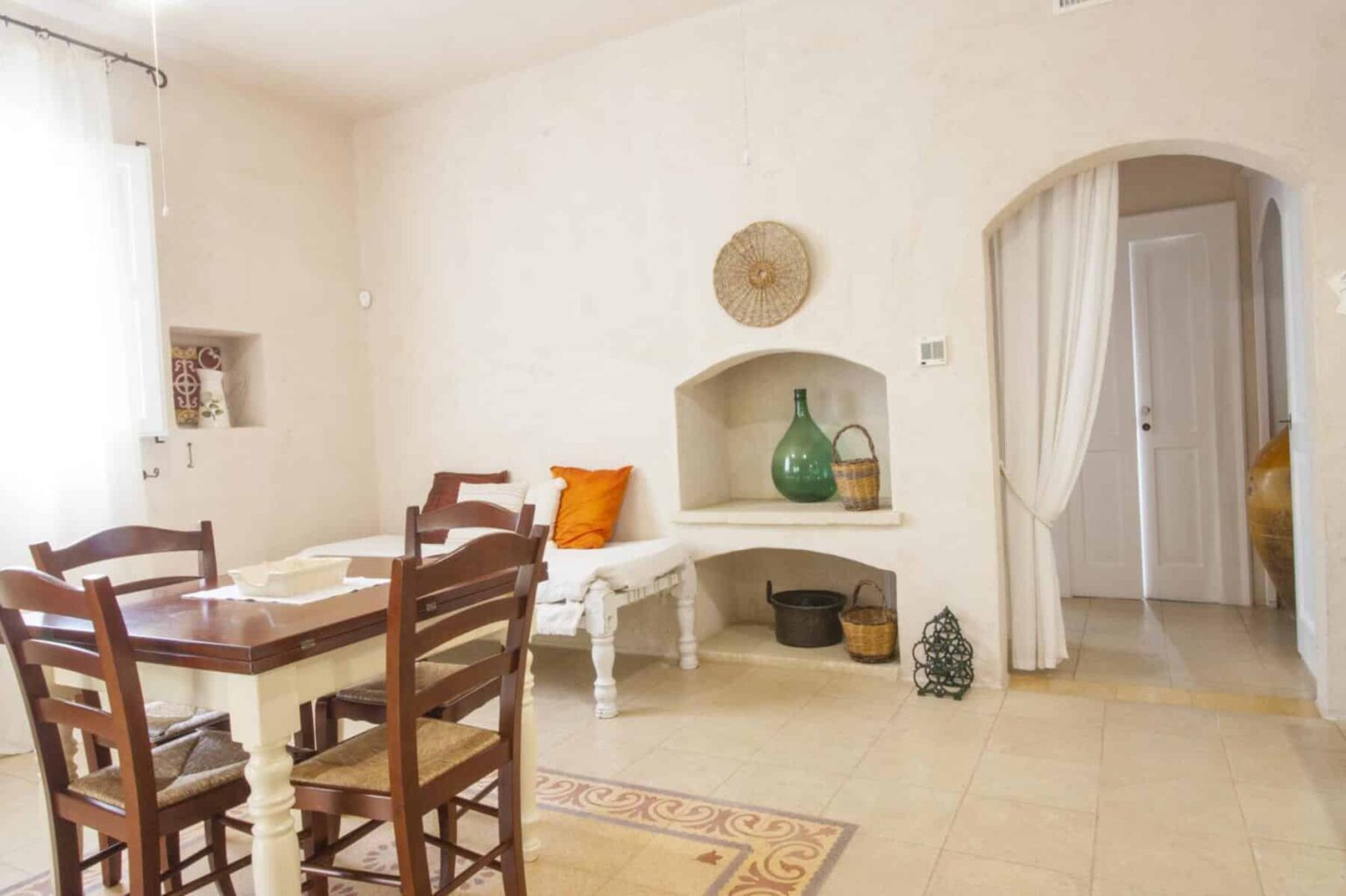 Cozy dining area featuring a wooden table with chairs, a decorative wall niche with a green vase, and warm-toned accents, illuminated by natural light from a window. The room has a neutral color palette and a welcoming atmosphere. | Only on SopranoVillas