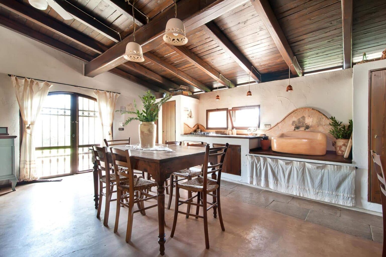 Cozy rustic kitchen and dining area with wooden beams, a large wooden table surrounded by chairs, and a stylish kitchen featuring a unique sink and natural light from windows. Decorative plants and soft curtains enhance the inviting atmosphere. | Only on SopranoVillas