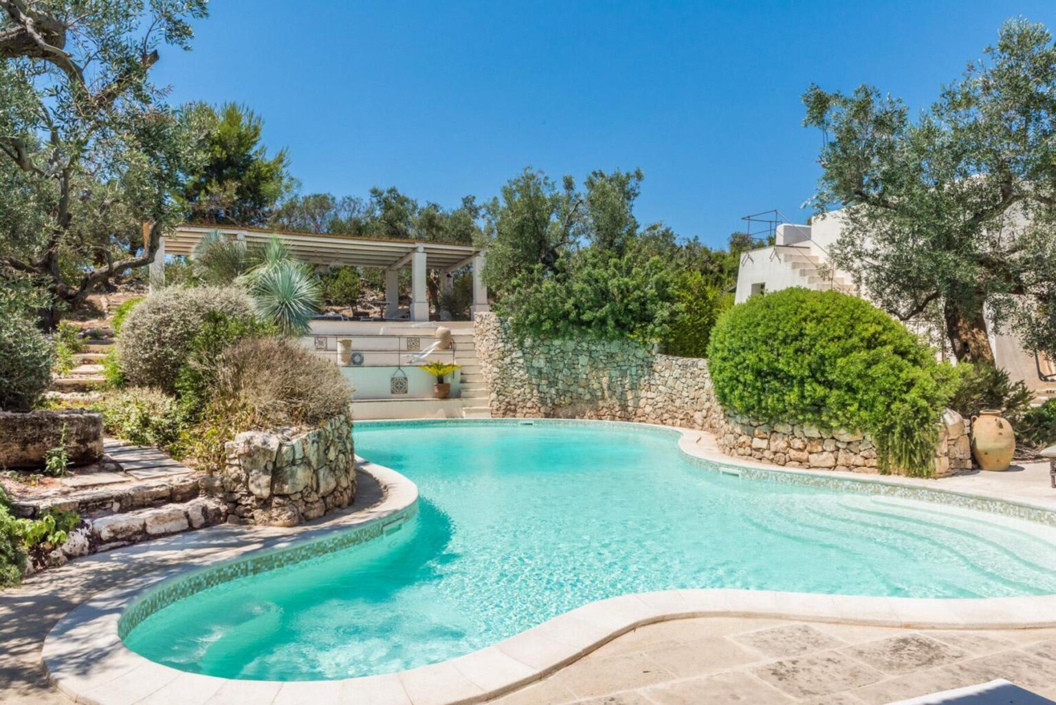 Serene outdoor swimming pool surrounded by lush greenery and stone walls, with a pergola and steps leading to a hillside, ideal for relaxation and leisure in a tranquil setting. | Only on SopranoVillas