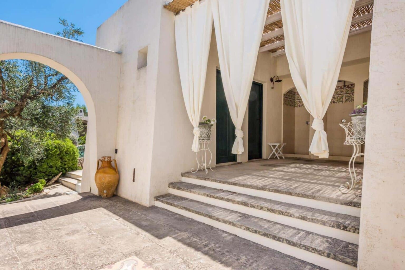 Elegant entrance of a Mediterranean-style villa featuring white draped curtains, stone steps, and a terracotta pot, surrounded by lush greenery and bright blue skies. | Only on SopranoVillas