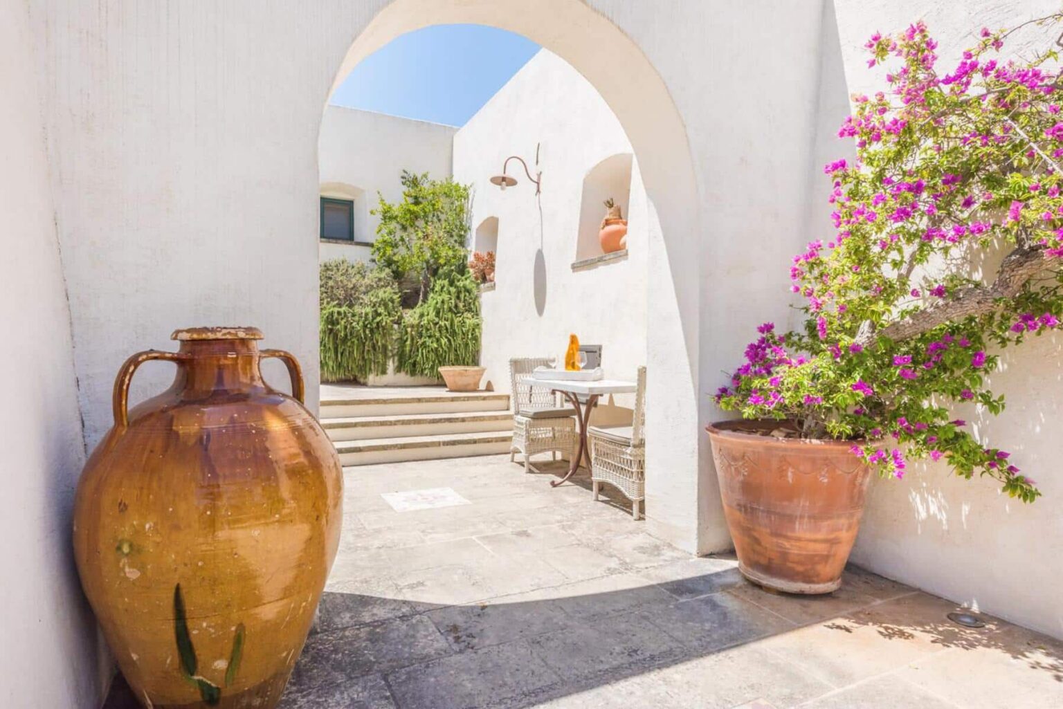 Charming courtyard featuring a large clay pot, a small table set for two, and vibrant bougainvillea against a backdrop of white walls and stairs, creating a serene outdoor space. | Only on SopranoVillas