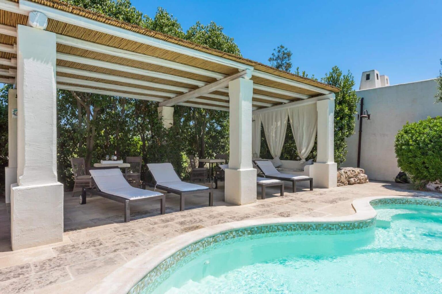 Relaxing outdoor patio with lounge chairs and a swimming pool, surrounded by lush greenery and a pergola for shade. Perfect for summer gatherings and leisure. | Only on SopranoVillas