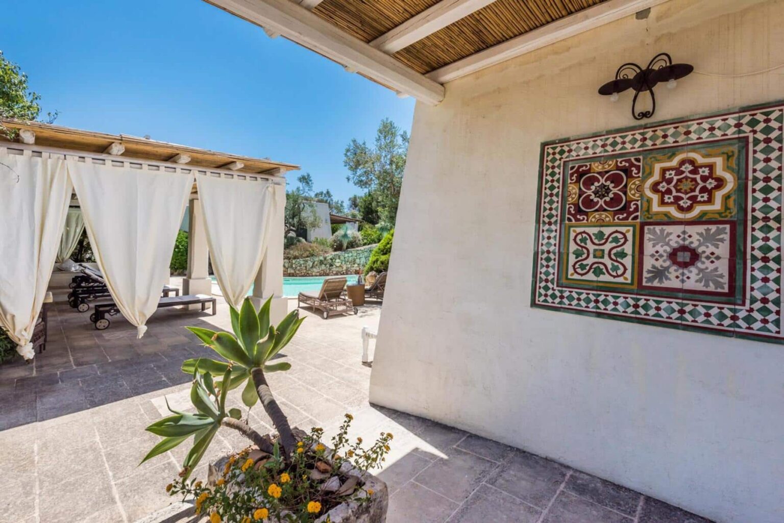 Outdoor patio area featuring a decorative tiled wall, potted plants, and a shaded seating area with draped curtains, overlooking a pool in a sunny garden setting. | Only on SopranoVillas