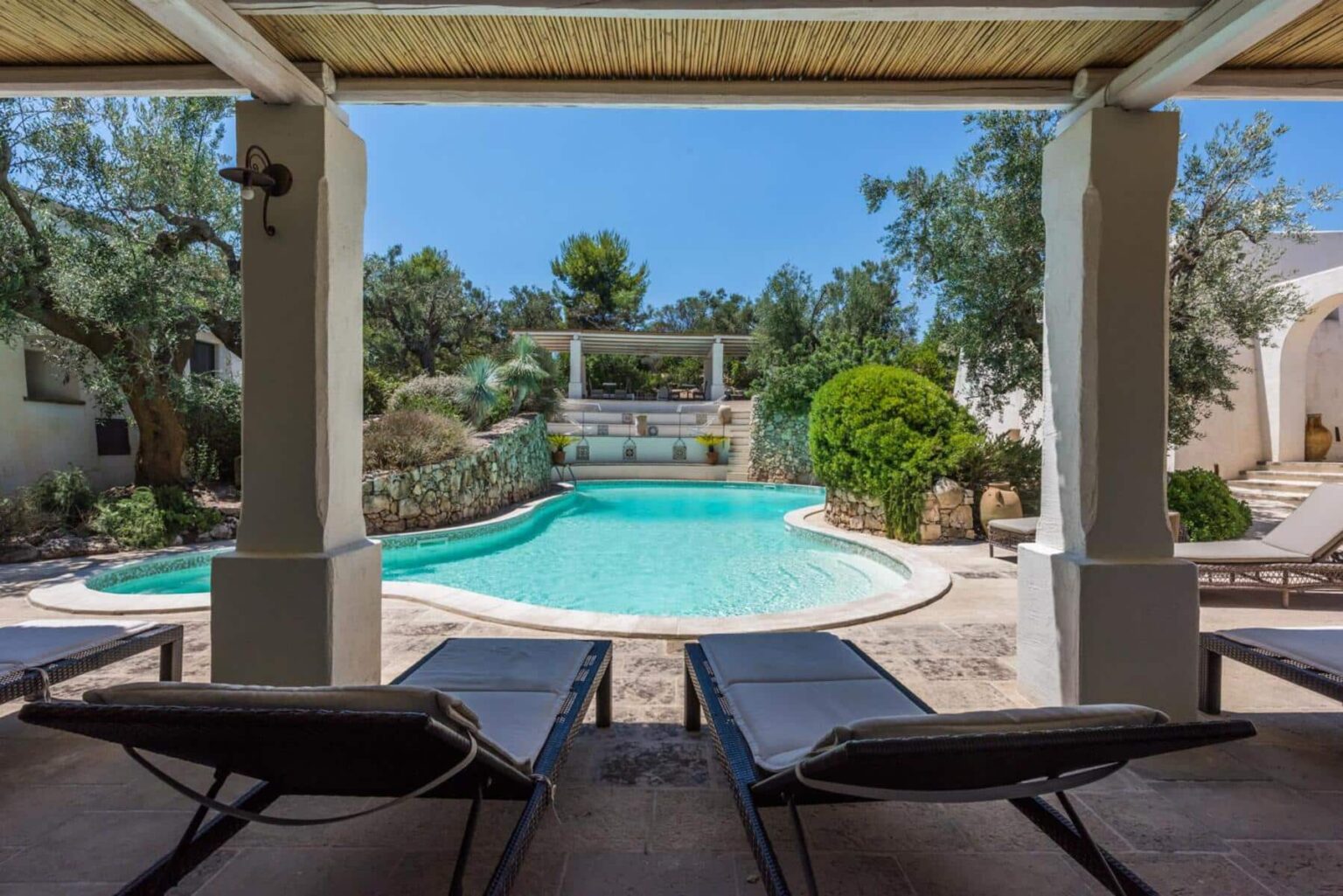 Relaxing outdoor space featuring lounge chairs by a sparkling swimming pool surrounded by lush greenery and stone landscaping under a clear blue sky. | Only on SopranoVillas