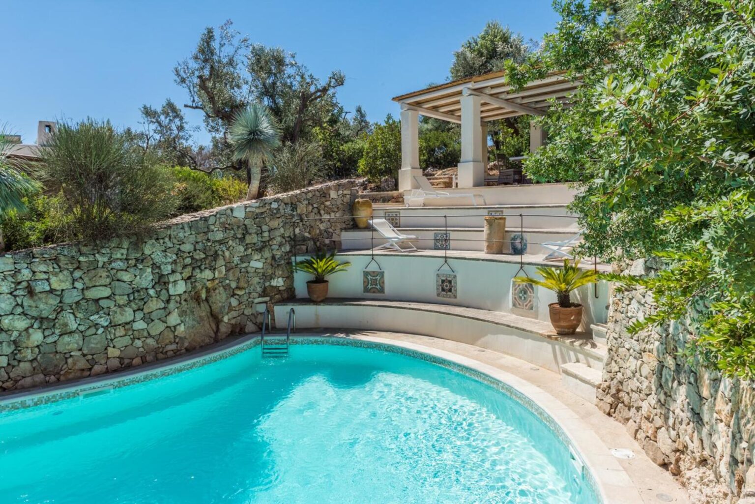 Serene outdoor swimming pool surrounded by natural stone walls and lush greenery, featuring a staircase leading to a shaded terrace with comfortable seating and decorative planters. Ideal for relaxation and outdoor gatherings in a tranquil setting. | Only on SopranoVillas