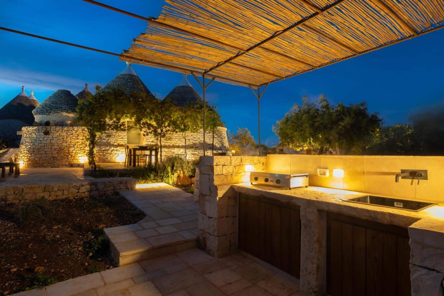 Outdoor kitchen area featuring a stone countertop and modern grill, set against traditional trulli houses in a serene evening atmosphere with ambient lighting and lush greenery. | Only on SopranoVillas