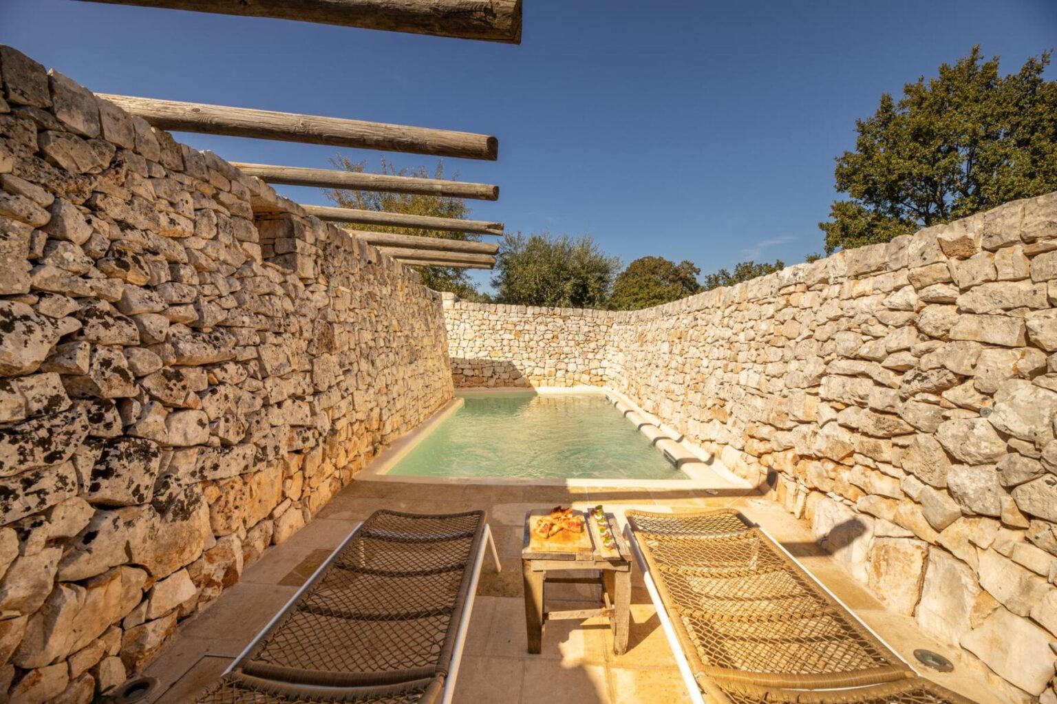 Serene outdoor pool area featuring natural stone walls, wooden beams, and comfortable lounge chairs, surrounded by lush greenery under a clear blue sky. Ideal for relaxation and leisure. | Only on SopranoVillas