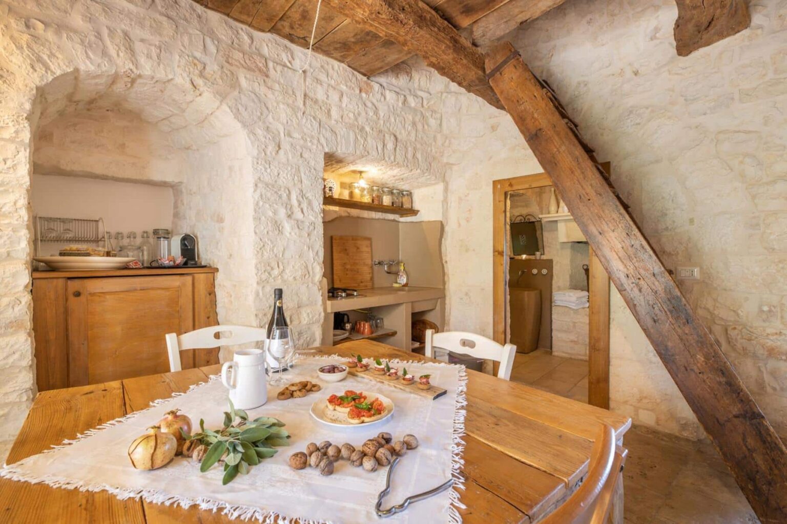 Charming rustic kitchen interior featuring a wooden dining table set with a variety of appetizers, including a plate of bruschetta, fruits, and nuts. The stone walls and wooden beams create a warm, inviting atmosphere, while a wine bottle and glasses add a touch of elegance. A cozy kitchen area is visible in the background, enhancing the homey feel. | Only on SopranoVillas
