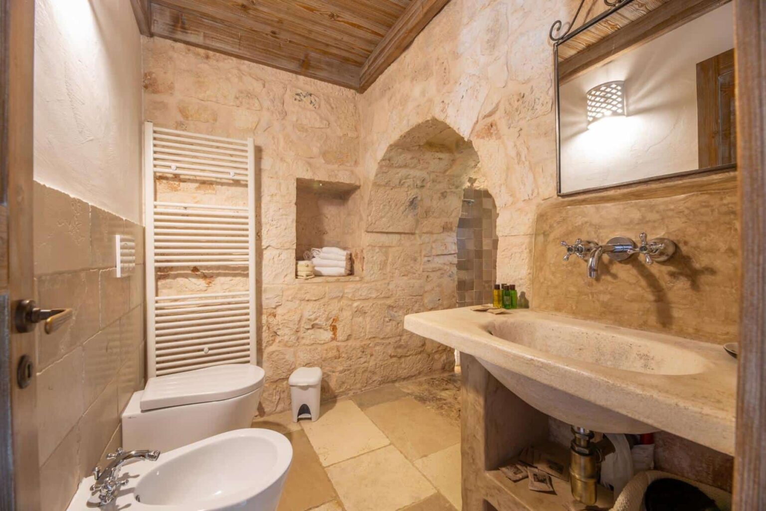 Modern bathroom with natural stone walls, featuring a sleek sink, bidet, and toilet. The space includes a towel rack and a shower area, complemented by wooden accents and elegant lighting. | Only on SopranoVillas