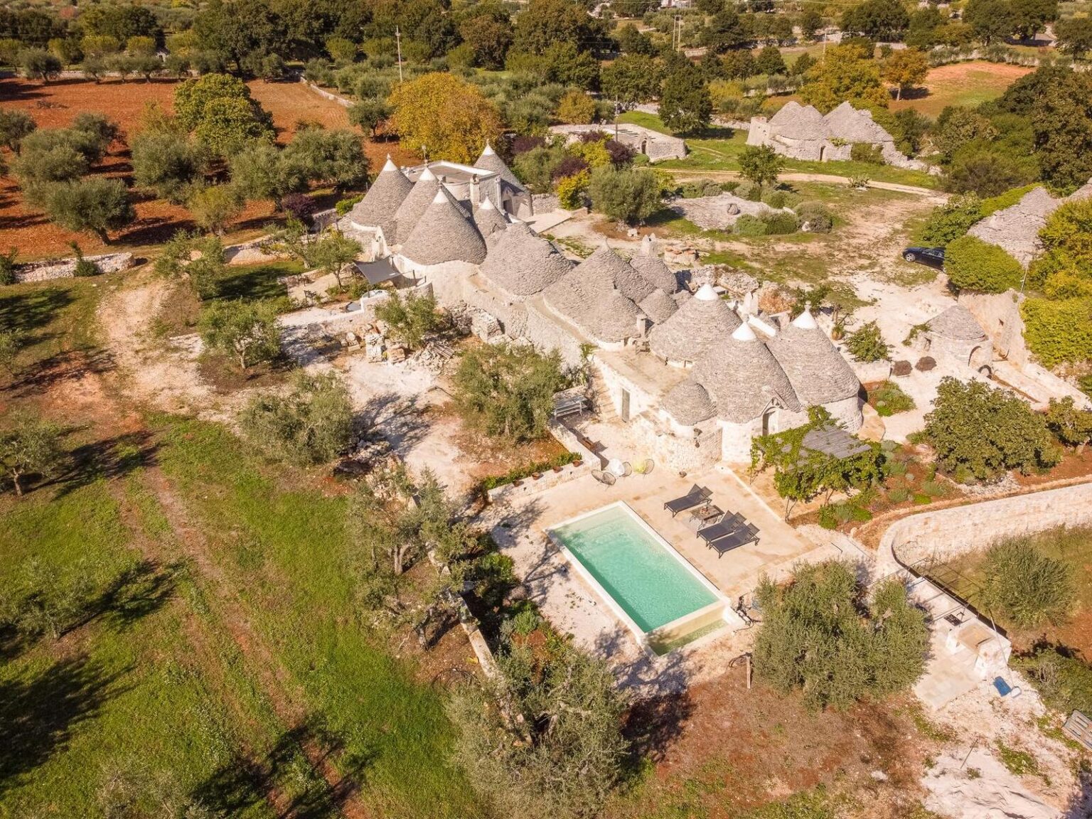 Aerial view of a charming rural villa featuring traditional trulli architecture, surrounded by olive trees and lush greenery, with a serene swimming pool in the foreground. | Only on SopranoVillas