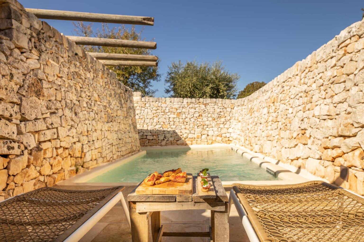 Serene outdoor pool area surrounded by rustic stone walls, featuring two lounge chairs and a wooden table with a gourmet meal. Ideal for relaxation and dining in a tranquil setting. | Only on SopranoVillas