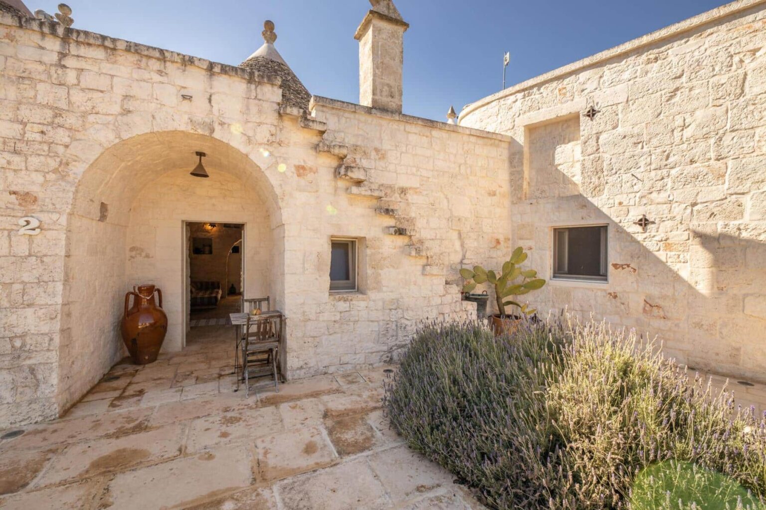 Charming stone cottage entrance featuring arched doorway, staircase, and terracotta pottery, surrounded by lavender and cacti in a serene outdoor setting. | Only on SopranoVillas