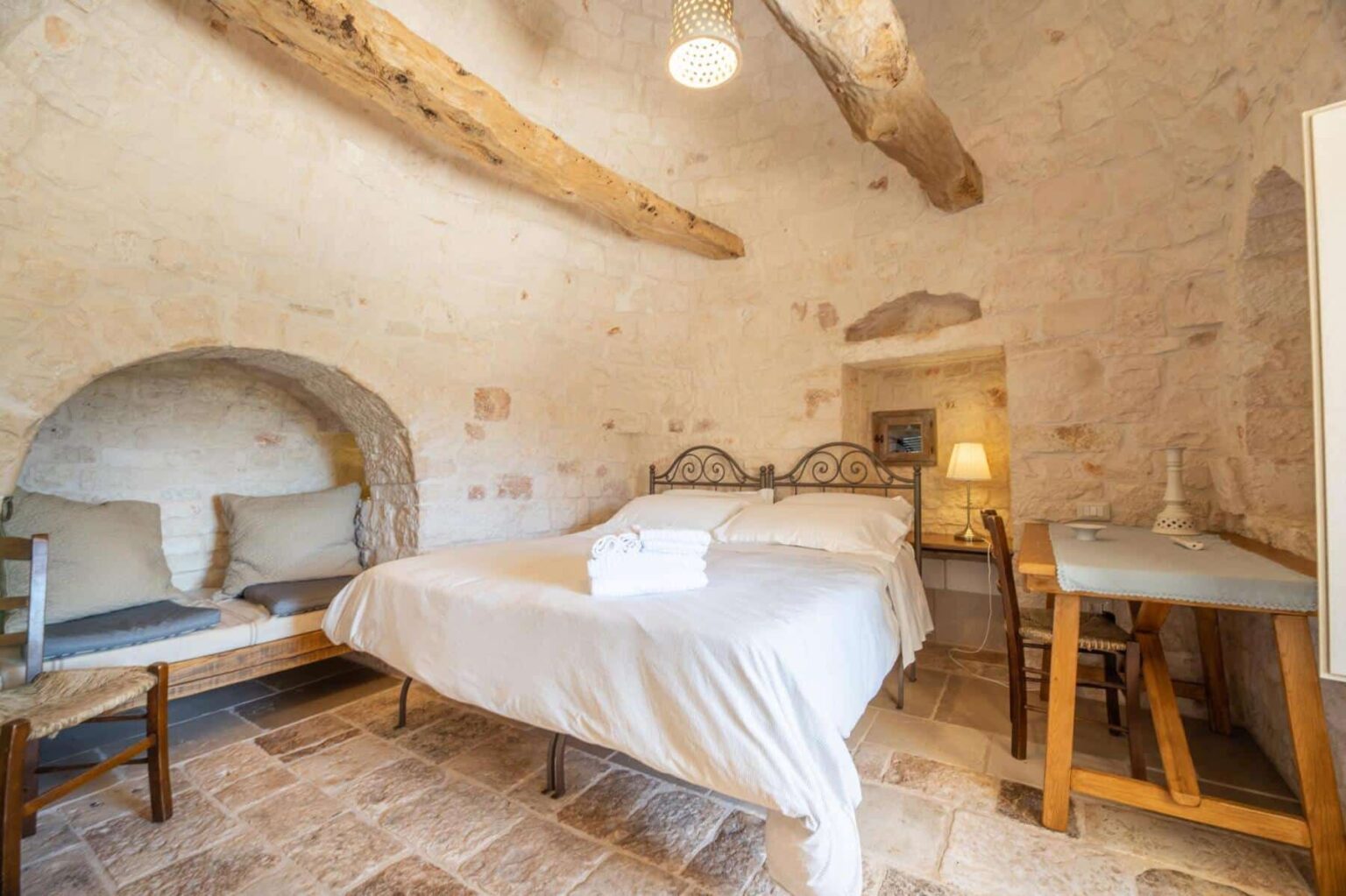 Cozy bedroom featuring a double bed with fresh linens, stone walls, wooden beams, and a small seating area. The room includes a bedside lamp and a wooden desk, creating a warm and inviting atmosphere. Ideal for a relaxing getaway. | Only on SopranoVillas