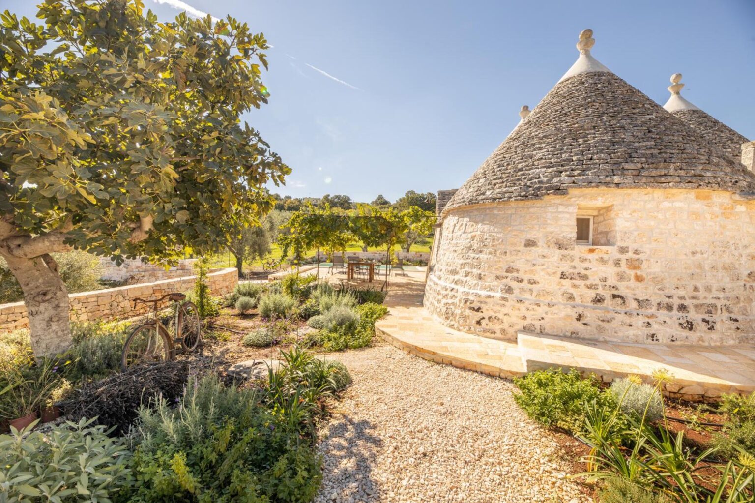 Charming trullo house surrounded by lush greenery and a gravel pathway, featuring a vintage bicycle and outdoor seating area in a picturesque garden setting. | Only on SopranoVillas